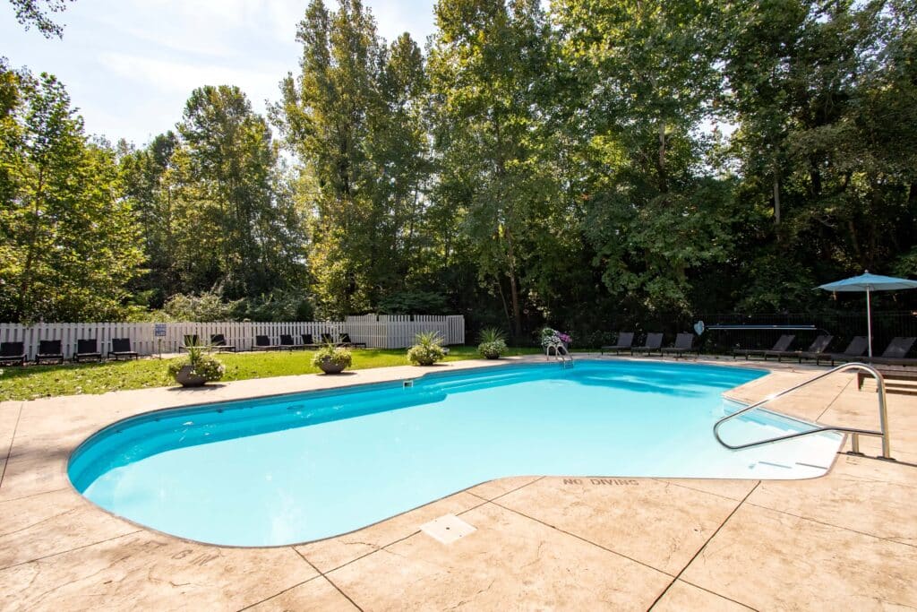 Outdoor swimming pool with clear water, surrounded by a concrete deck. Lounge chairs and umbrellas are on one side, with trees and greenery in the background.