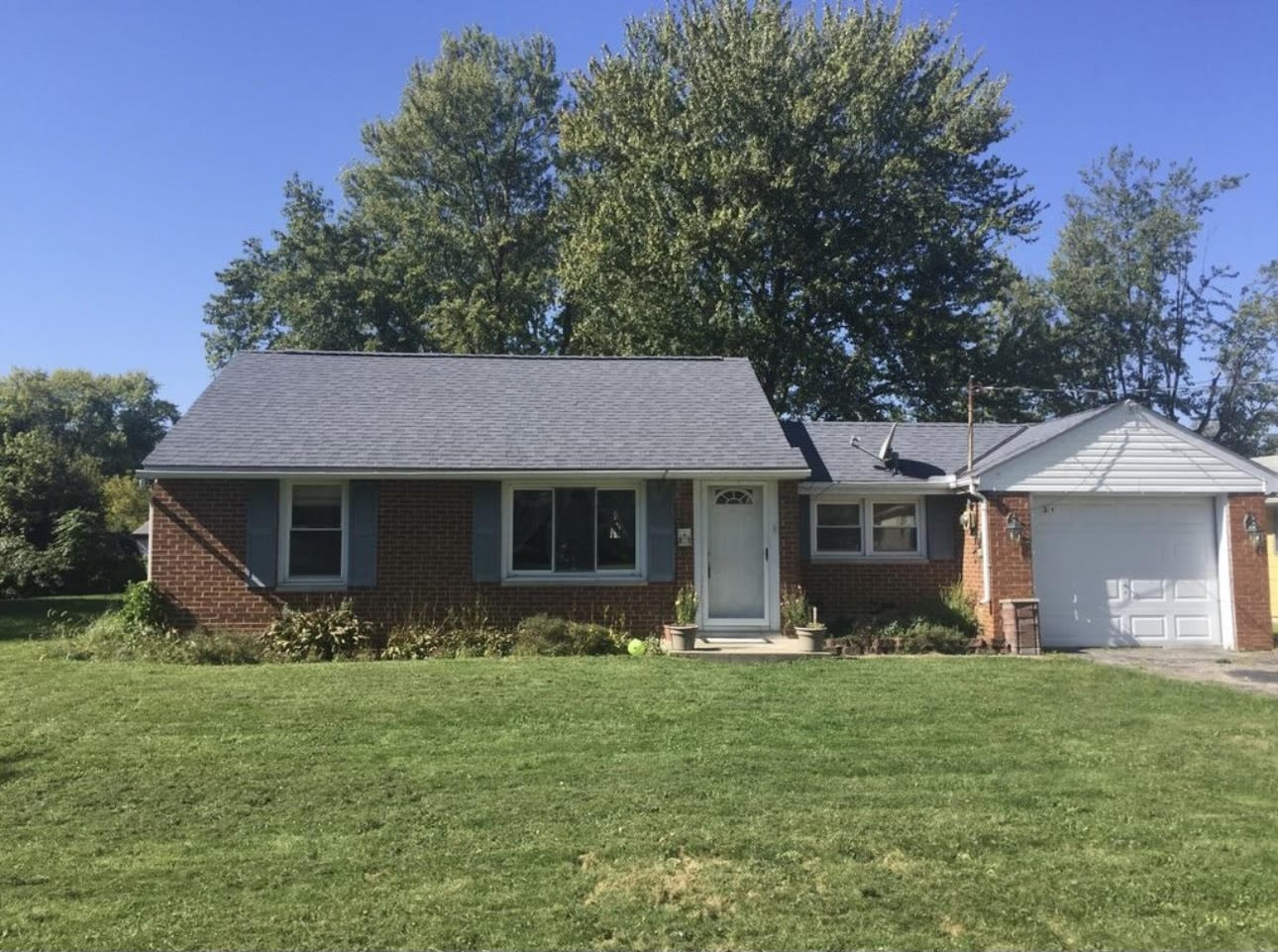 A brick single-story house with a gray roof, attached garage, and a small front porch. Surrounded by green grass and trees under a clear blue sky.