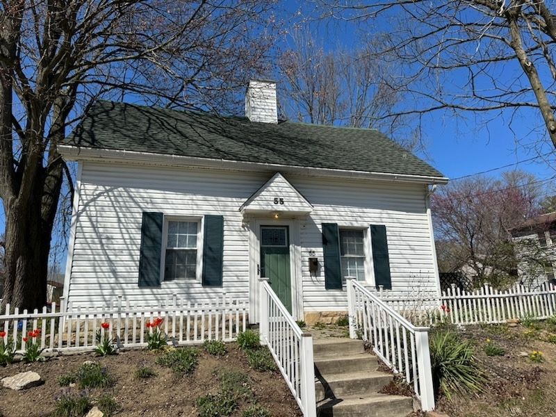 White house with green shutters and front door, chimney, and white picket fence. Red tulips and flowers line the path to the entrance. Leafless trees surround the house under a clear blue sky.