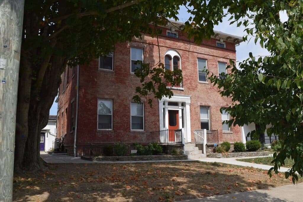 A three-story red brick building with arched windows and a white-trimmed entry door, surrounded by trees and a patchy lawn.