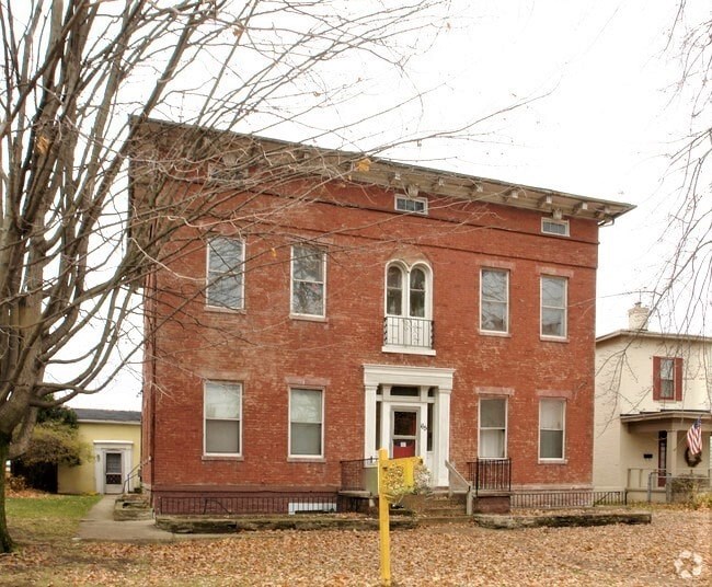 A two-story red brick building with several windows, a centered front door, and a small balcony, surrounded by leafless trees and fallen leaves on the ground.