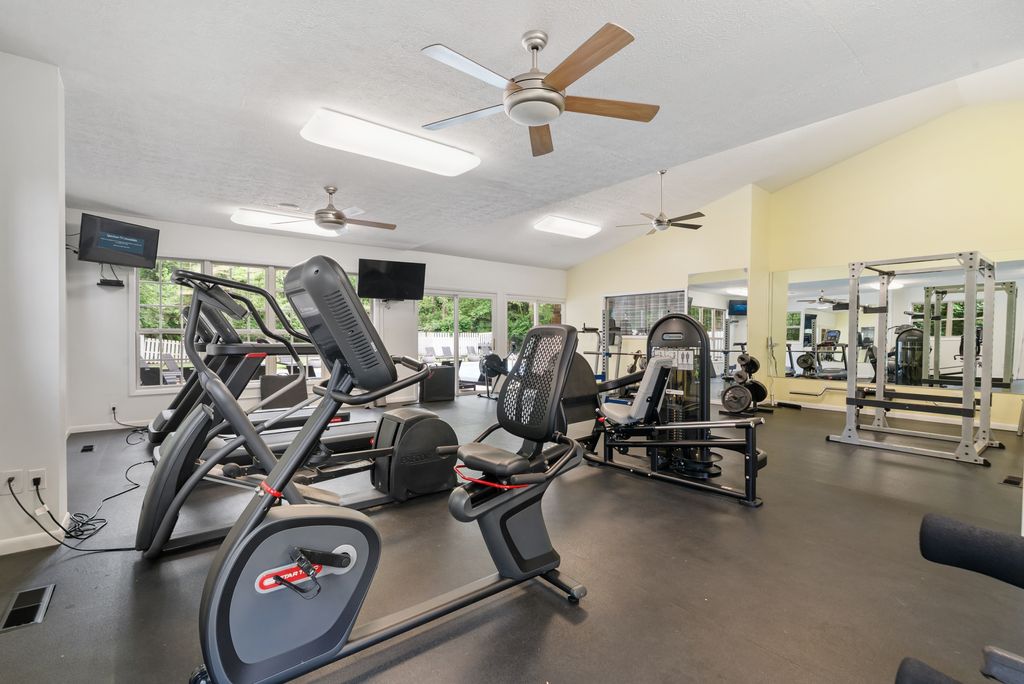 Modern gym with exercise bikes, treadmills, weight machines, and large windows letting in natural light; ceiling fans and mounted TVs are visible.