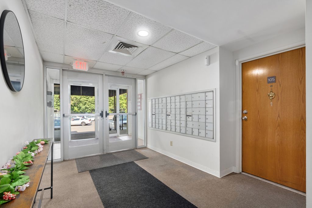 Apartment building lobby with mailboxes on the wall, a wooden door labeled 105, glass double entry doors, and a small table with decorative plants.