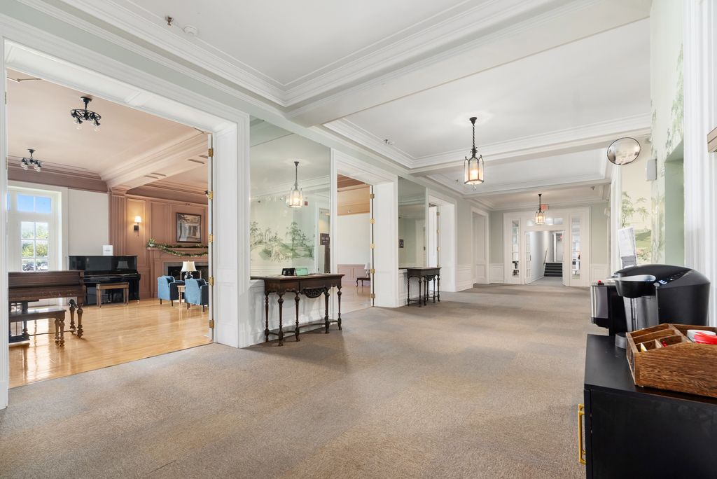 Spacious hallway with beige carpet, coffee station on the right, open rooms with pianos on the left, and pendant lighting throughout.