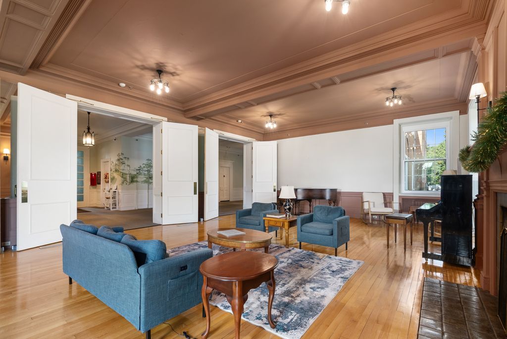 Spacious living room with wooden floors, blue sofas, a rug, a round wooden table, a piano, and large white doors leading to another room.