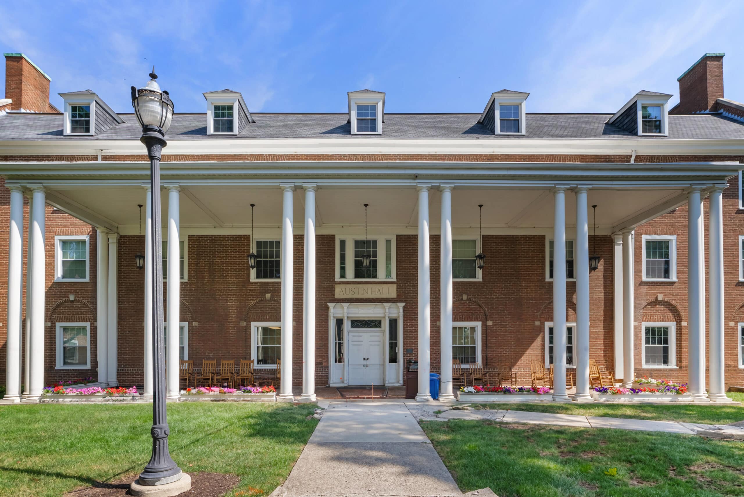 A brick building with white columns, multiple windows, and a front porch with chairs, labeled "Aston Hall." A lamp post and a walkway lead to the entrance.
