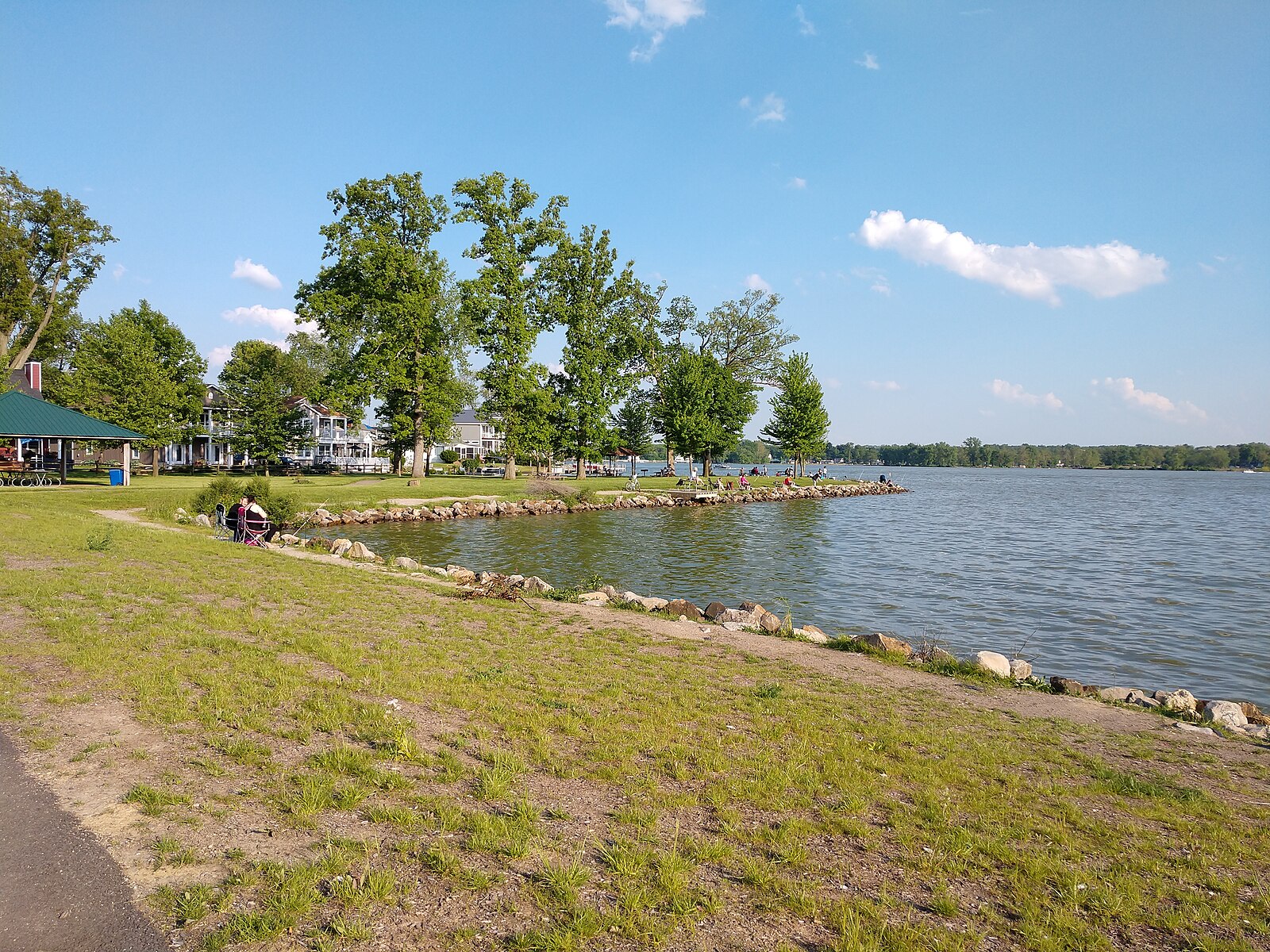 A lakeside scene with a grassy shore, trees, and a line of rocks bordering the water. People are relaxing and sitting by the edge of the lake under a clear sky.