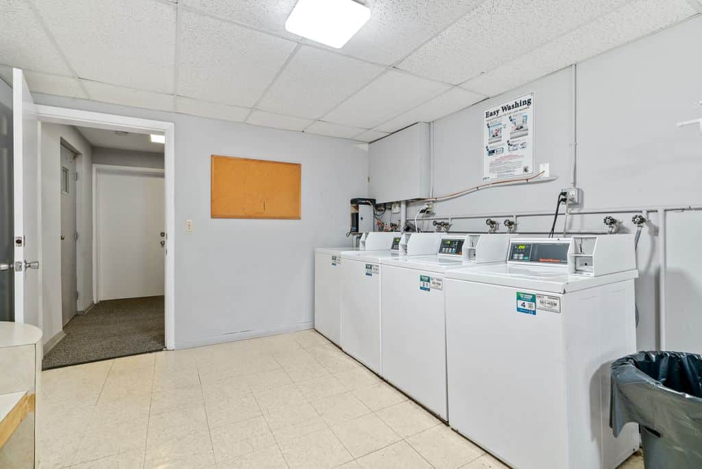 A laundry room with four white washing machines, a wall-mounted instruction sign, an empty bulletin board, and a trash can on a tiled floor.
