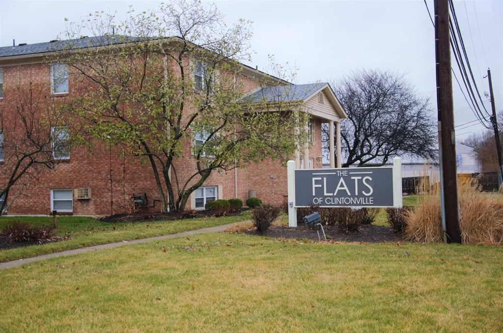 Brick apartment building with a sign reading "The Flats of Clintonville" on a grassy lawn. Trees and shrubs are in the front yard.