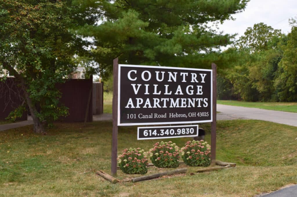Sign for Country Village Apartments at 101 Canal Road, Hebron, OH, with contact number 614.340.9830, situated outdoors with a background of trees and grass.