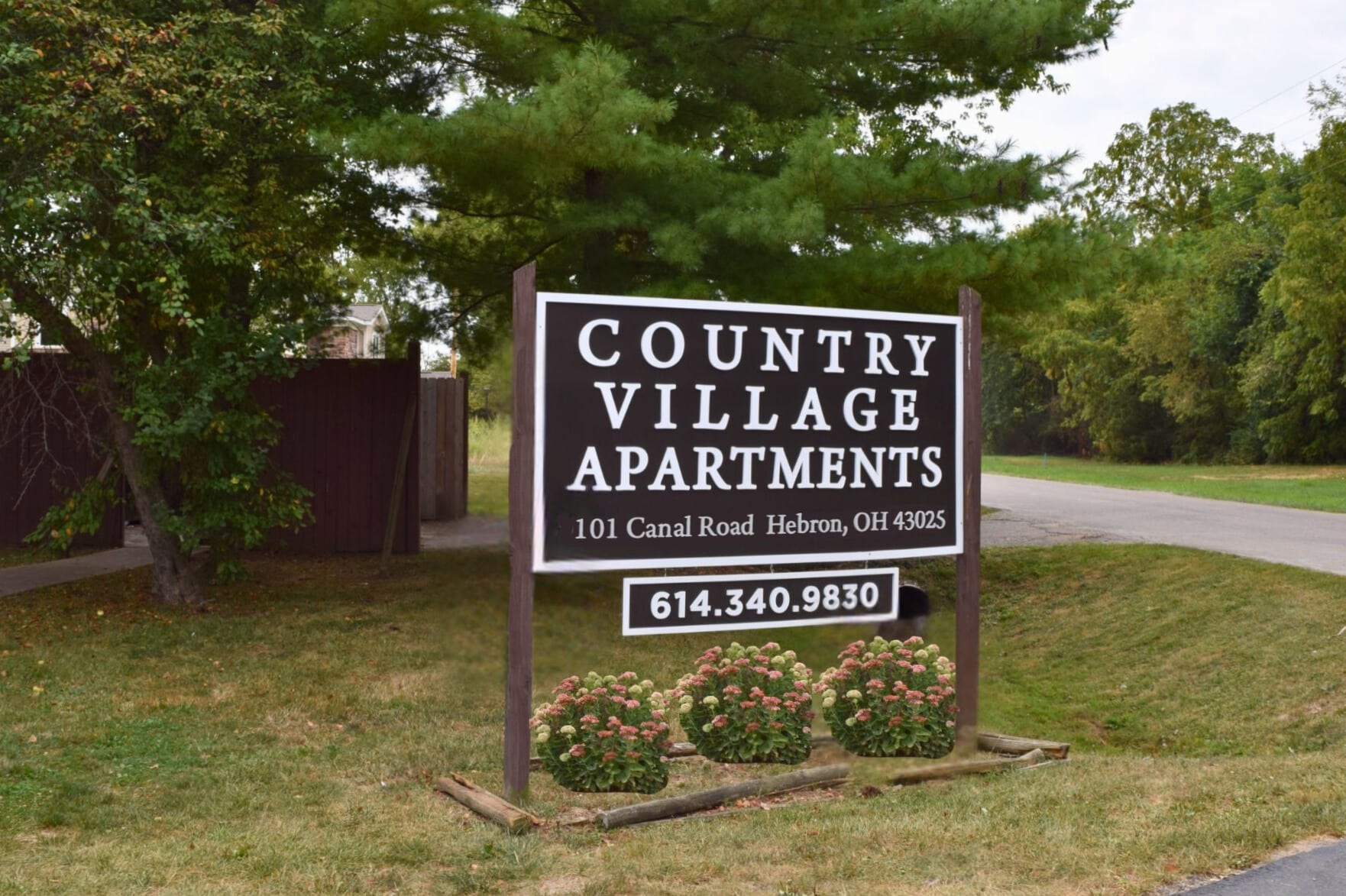Sign for Country Village Apartments at 101 Canal Road, Hebron, OH, with contact number 614.340.9830, situated outdoors with a background of trees and grass.