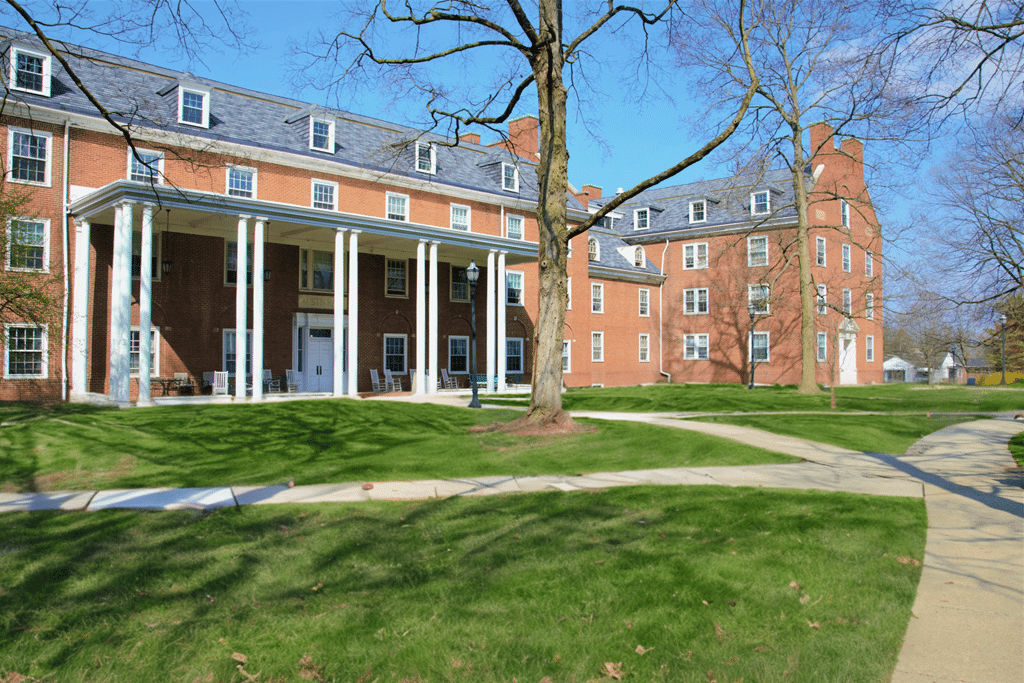 Brick building with white columns and multiple windows, surrounded by green grass and bare trees, under a clear blue sky. Sidewalks lead to and from the building's entrance.