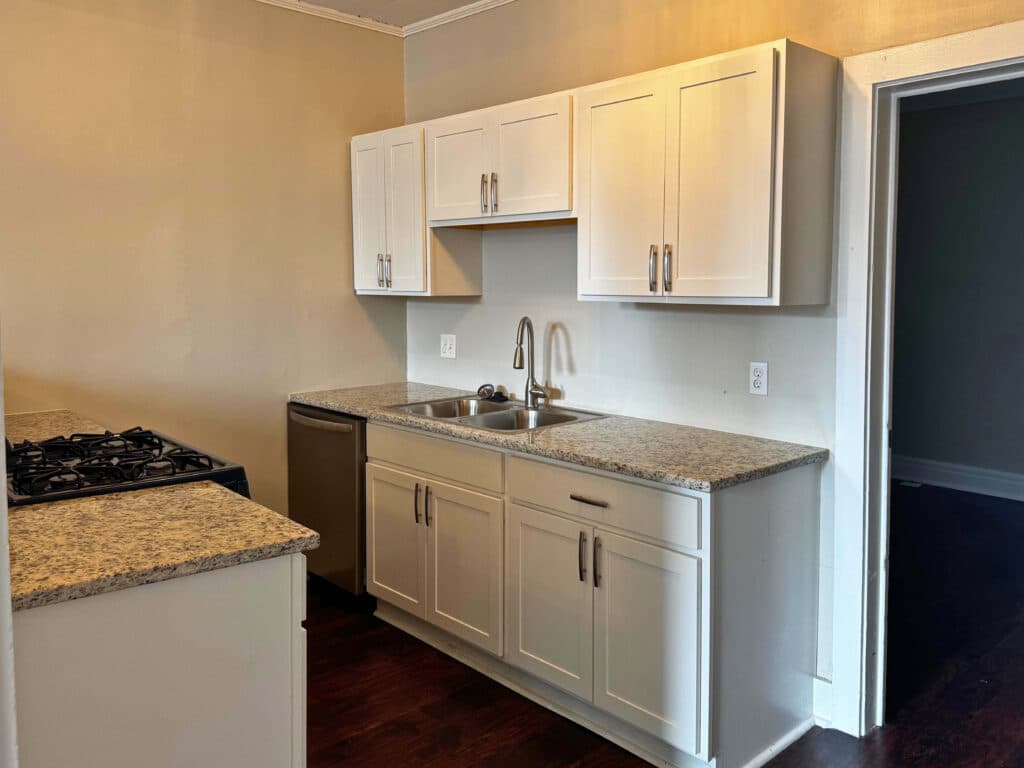 A kitchen with white cabinets, a stainless steel sink, and a granite countertop. Gas stovetop on the left. Brown flooring and beige walls.