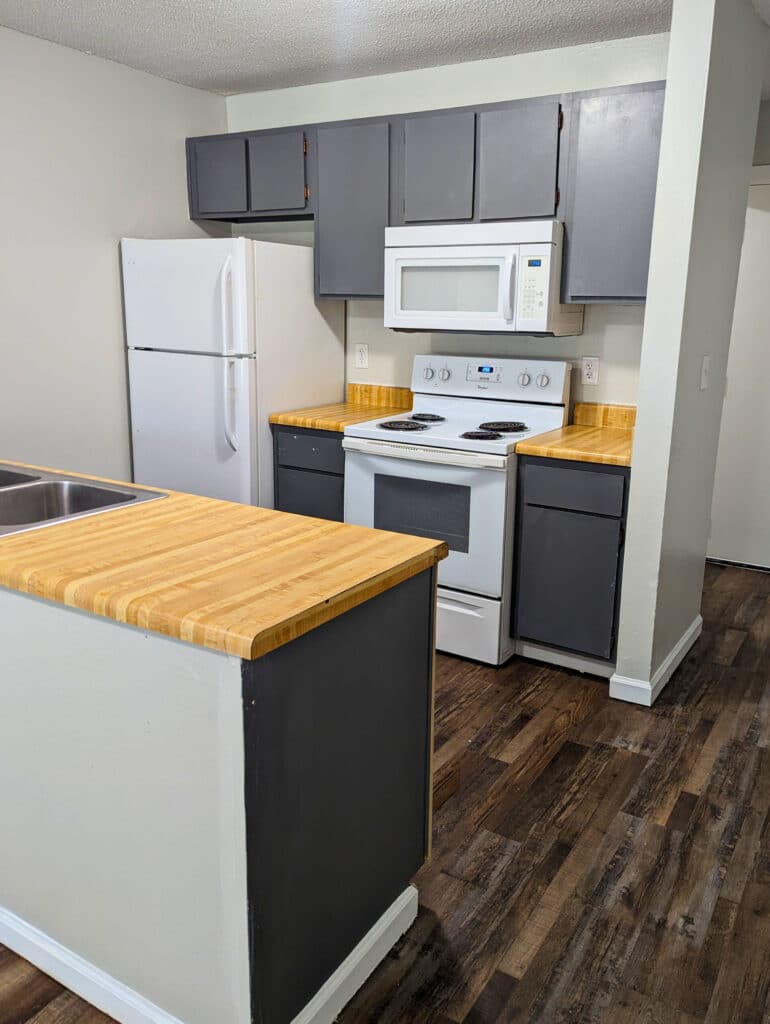 A small kitchen with gray cabinets, wood countertops, a white refrigerator, stove, microwave, and a kitchen island, featuring dark wood flooring.