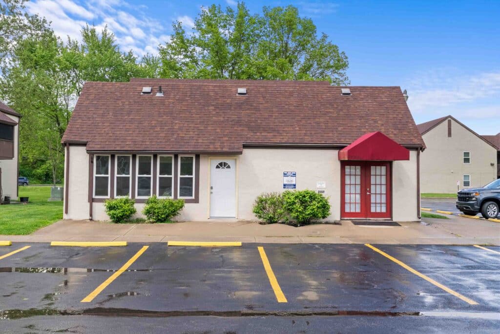 One-story building with a brown roof, white walls, red awning over a glass door, white entry door, and adjacent parking spaces on a cloudy day.