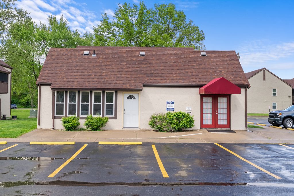 Single-story building with tan stucco walls, brown shingled roof, white and red doors, front bushes, and an adjacent parking lot with yellow-lined spaces.