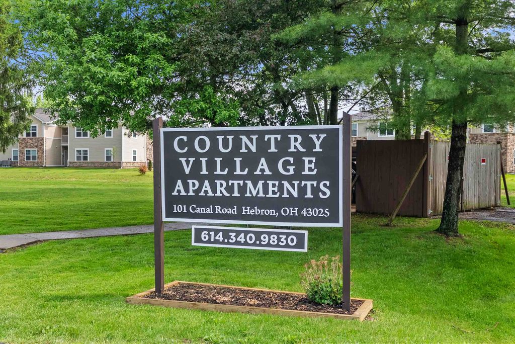 A sign reads "Country Village Apartments, 101 Canal Road, Hebron, OH 43025, 614.340.9830" in front of a grassy area with trees and apartment buildings in the background.