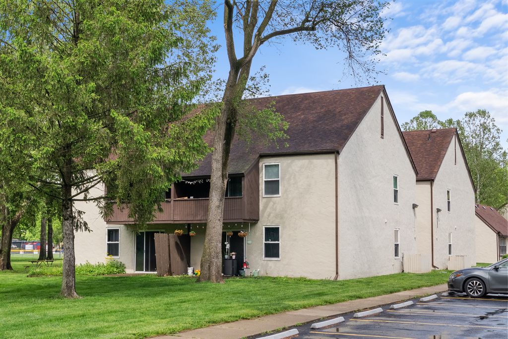 Two-story beige apartment building with brown roof, outdoor balcony, and a grassy yard, adjacent to a parking lot with a single parked car. Trees and blue sky in the background.