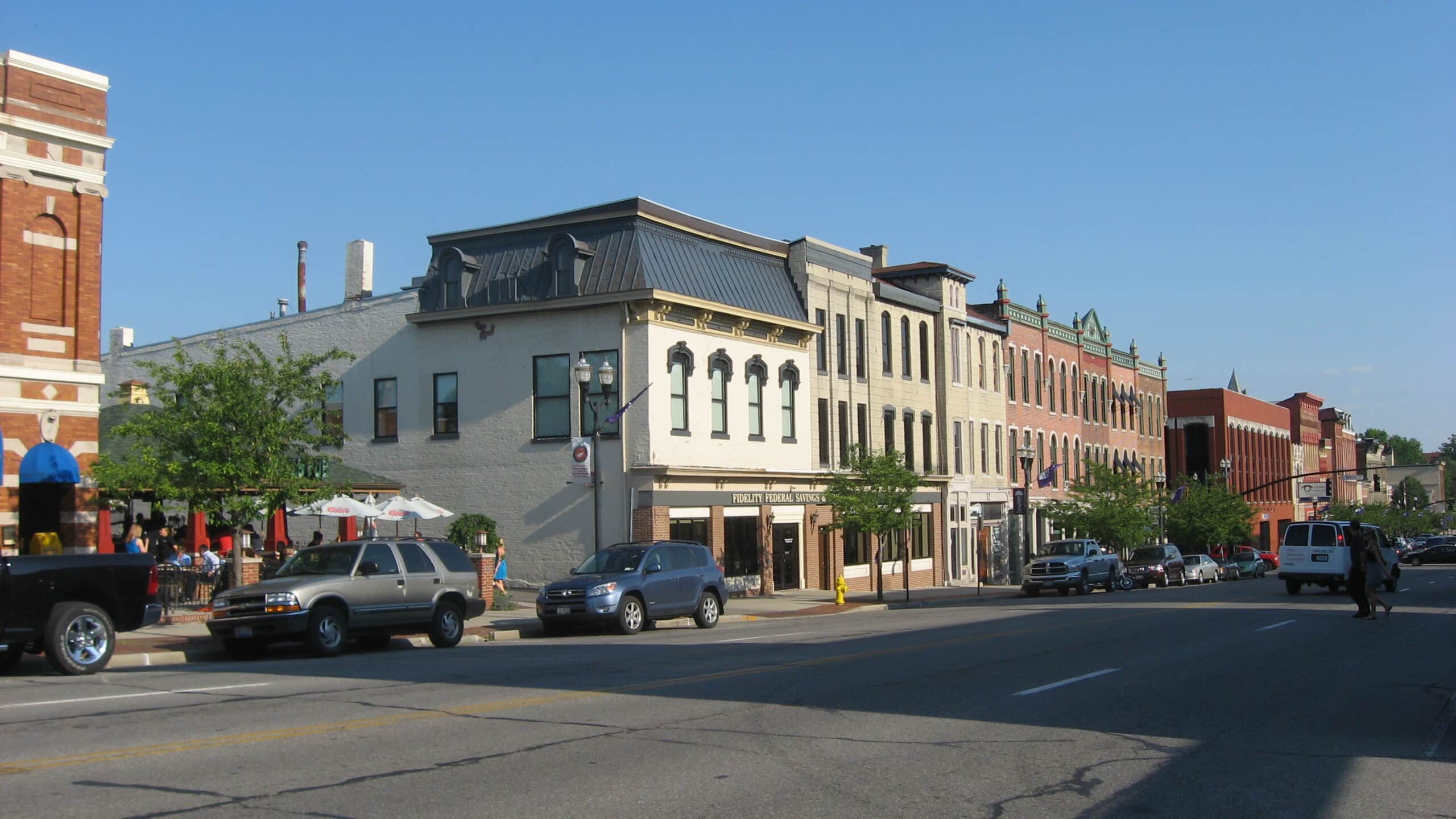 Street view of a small town with brick buildings, varied architecture, and parked cars along the road under a clear blue sky.