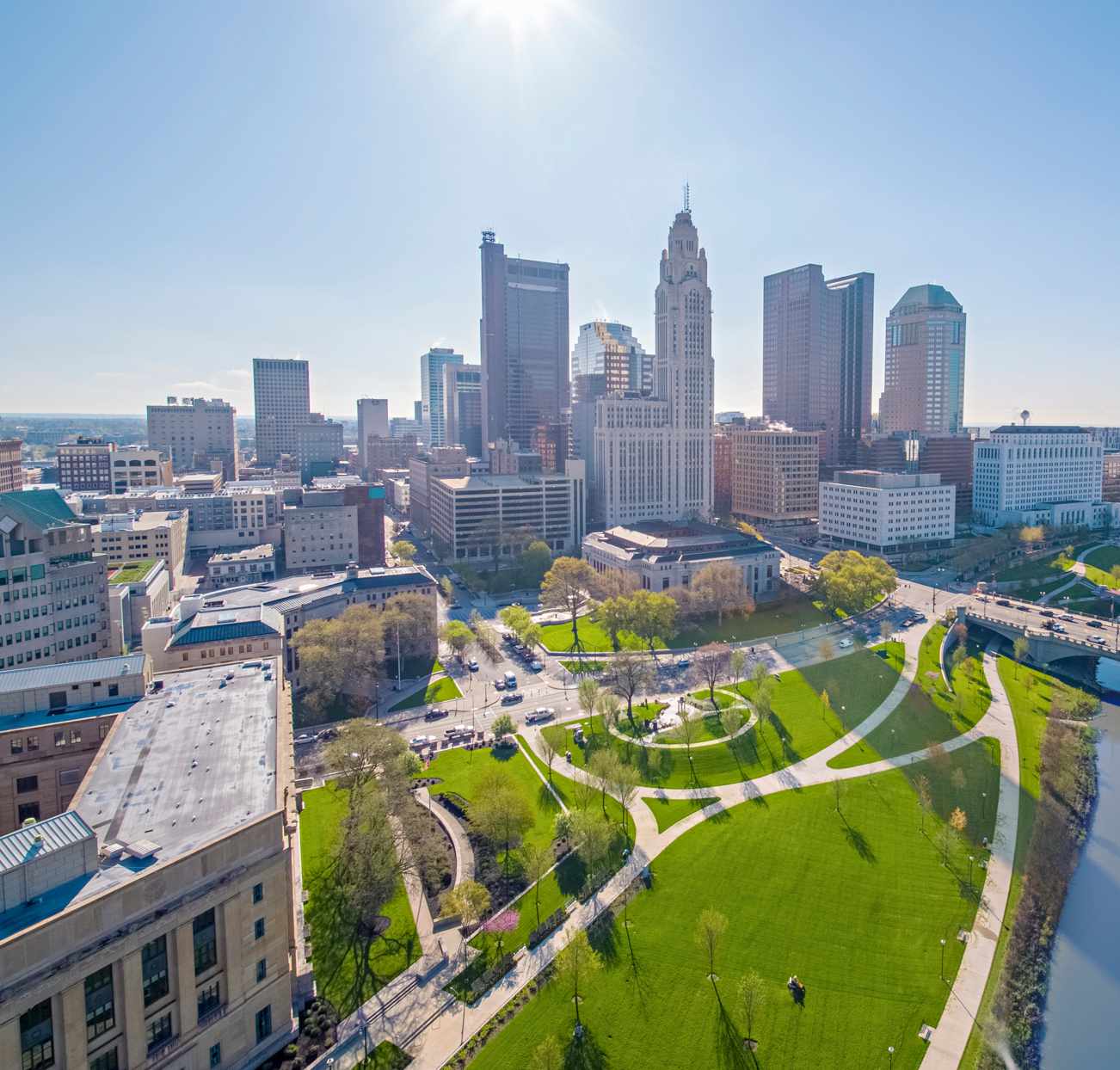 Aerial view of a city skyline with tall buildings, a green park, and a river on a sunny day.
