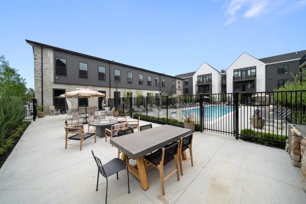 Outdoor patio area with tables and chairs next to a fenced swimming pool in a modern apartment complex under a clear blue sky.